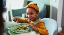 child smiling while eating food off a plate with a spoon