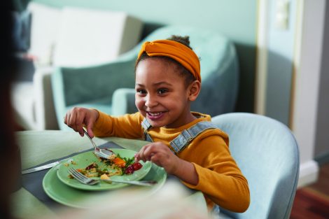 child smiling while eating food off a plate with a spoon