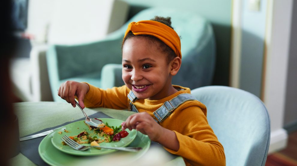 child smiling while eating food off a plate with a spoon