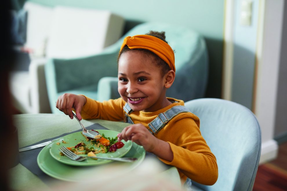 child smiling while eating food off a plate with a spoon