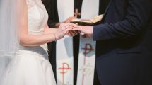 Bride slipping wedding ring onto her partner's finger in front of a priest.