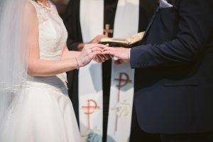 Bride slipping wedding ring onto her partner's finger in front of a priest.