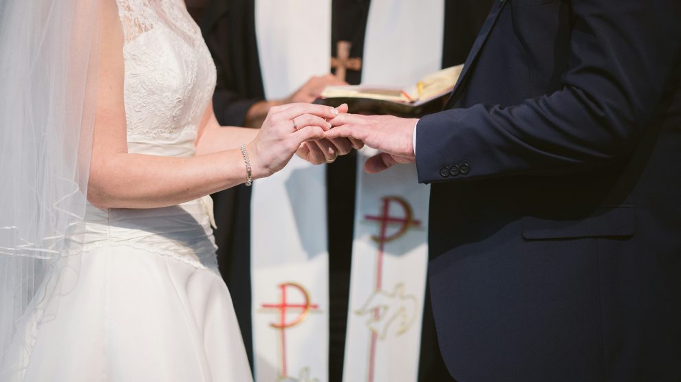 Bride slipping wedding ring onto her partner's finger in front of a priest.