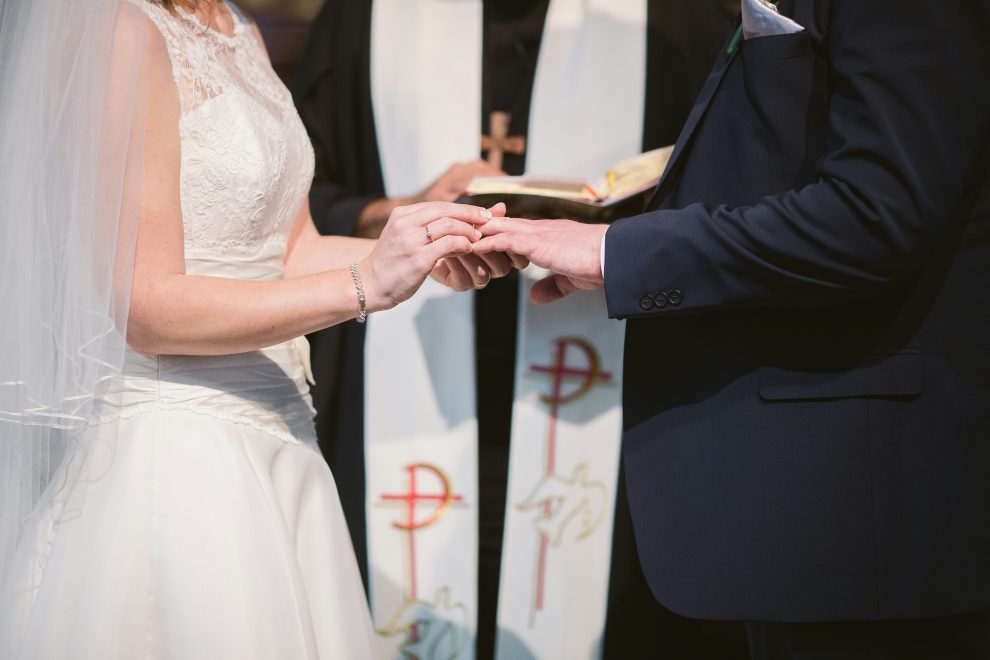 Bride slipping wedding ring onto her partner's finger in front of a priest.