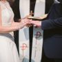 Bride slipping wedding ring onto her partner's finger in front of a priest.